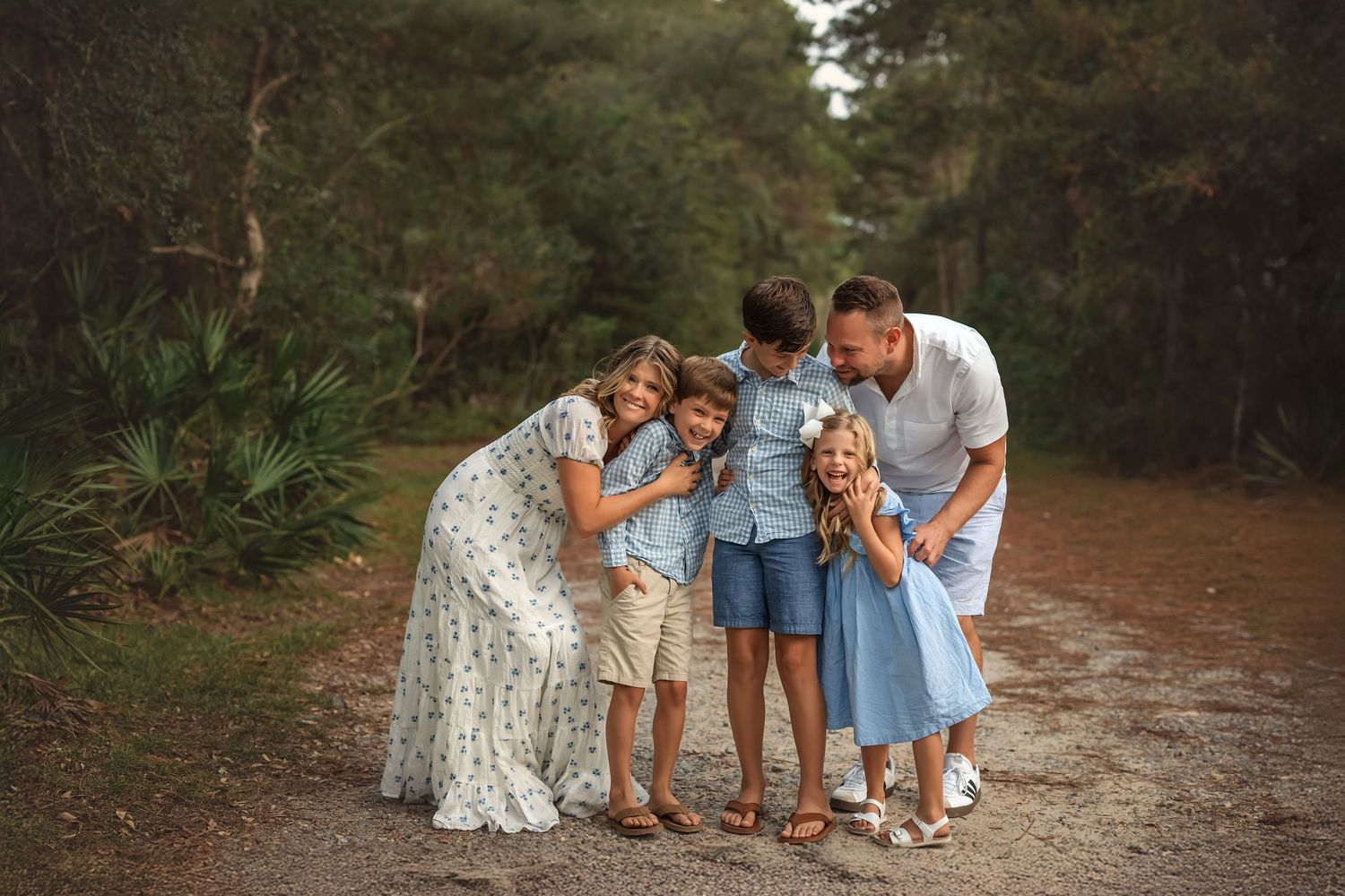 Family of five sharing a joyful moment on a forest path, dressed in coordinating blue and white outfits.