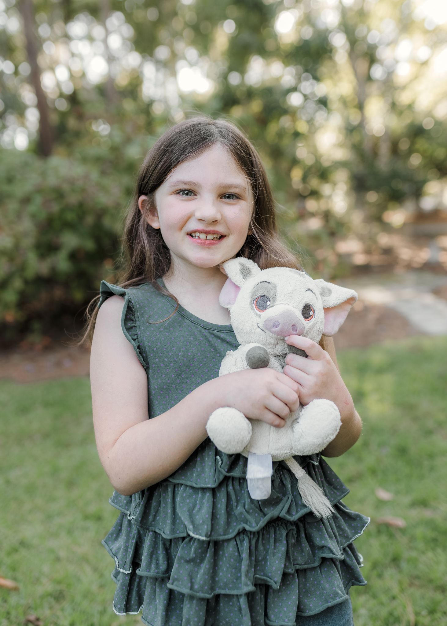 Young girl in gray ruffled dress smiling outdoors while hugging a white pig stuffed animal toy.