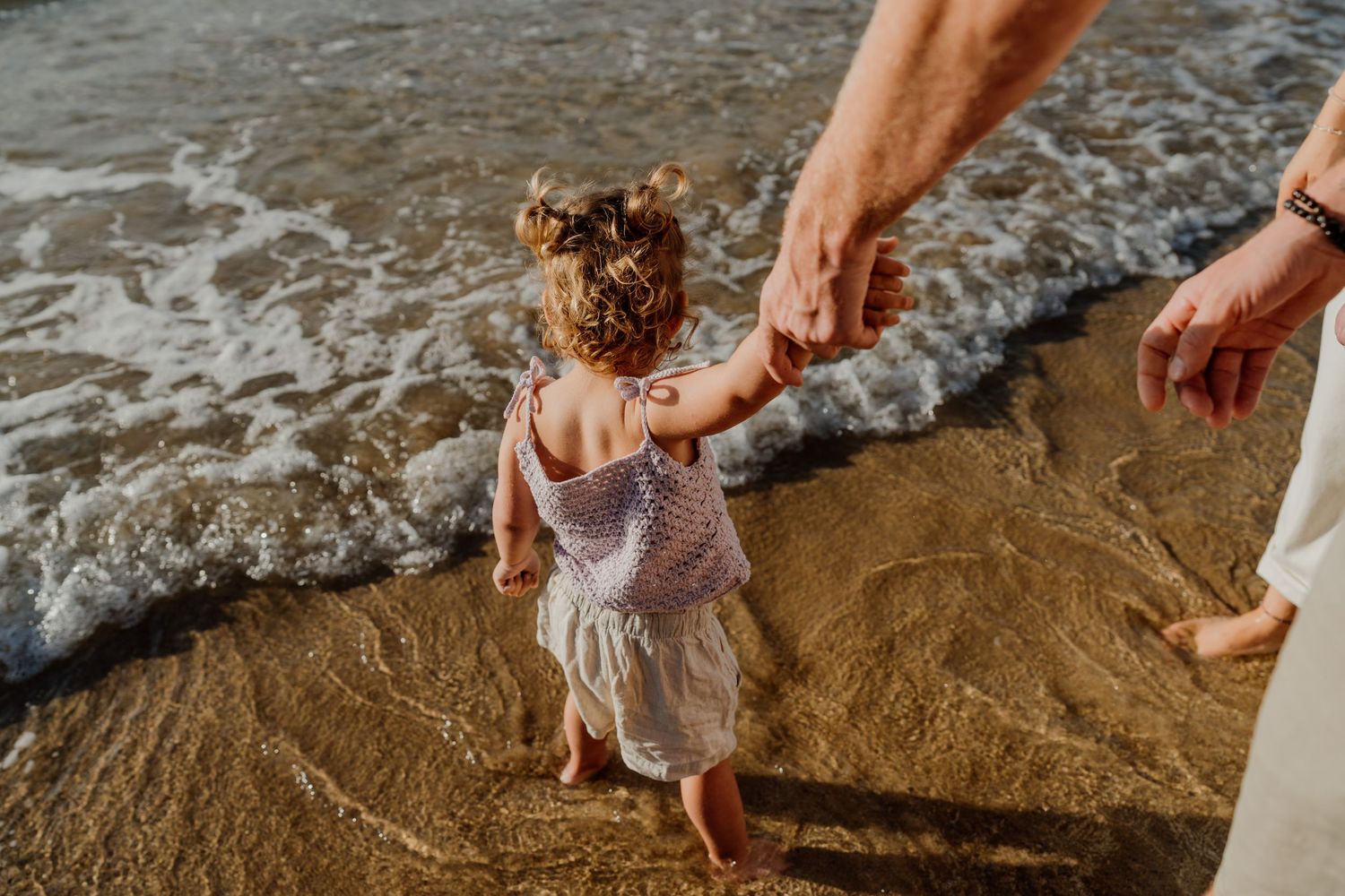 A toddler girl walks barefoot on a sandy beach holding an adult's hand as gentle waves wash ashore.