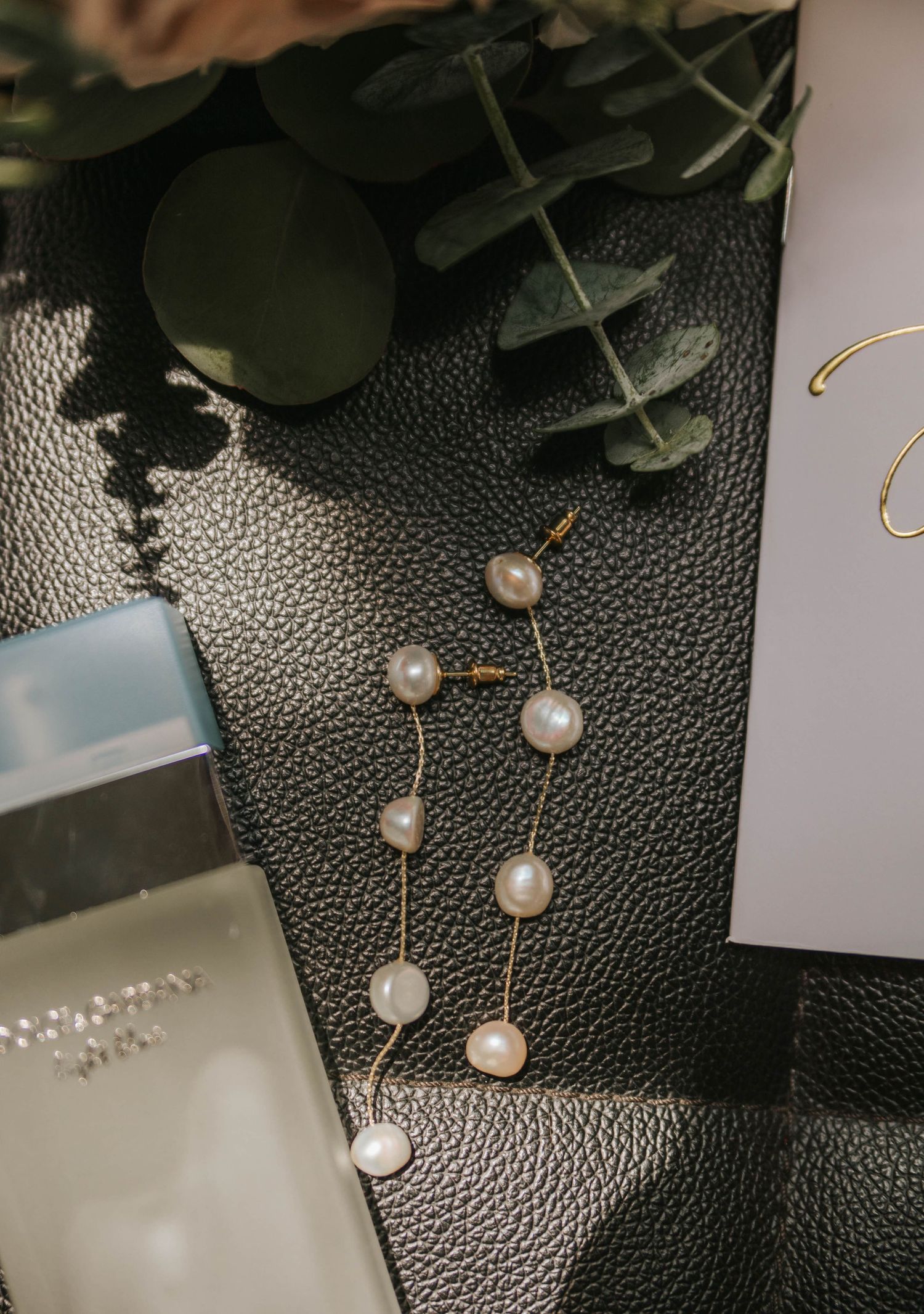 Gold chain earrings with white freshwater pearls displayed on black leather surface with greenery and elegant stationery.
