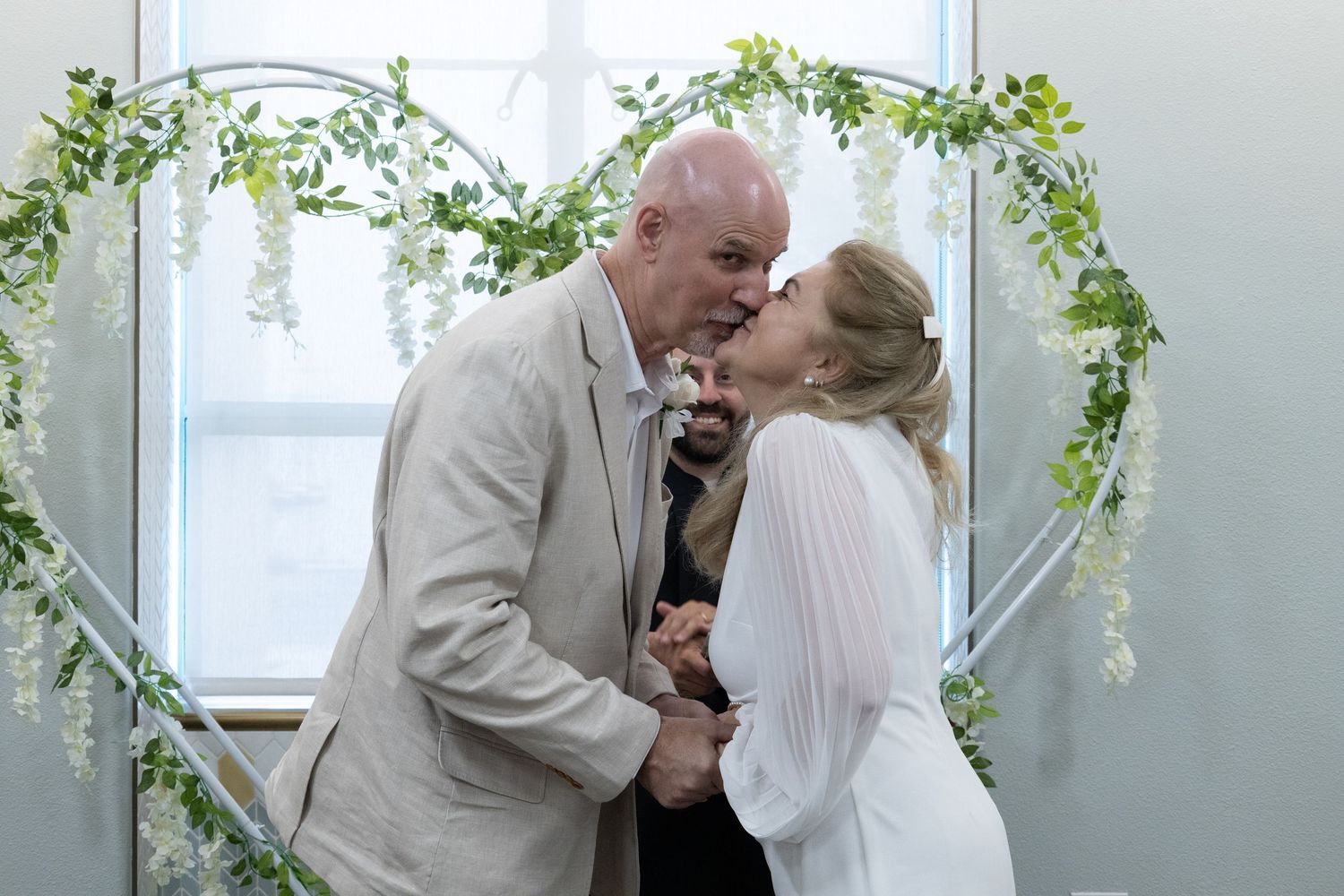 Couple sharing a kiss at wedding ceremony under floral heart arch with greenery and white flowers.