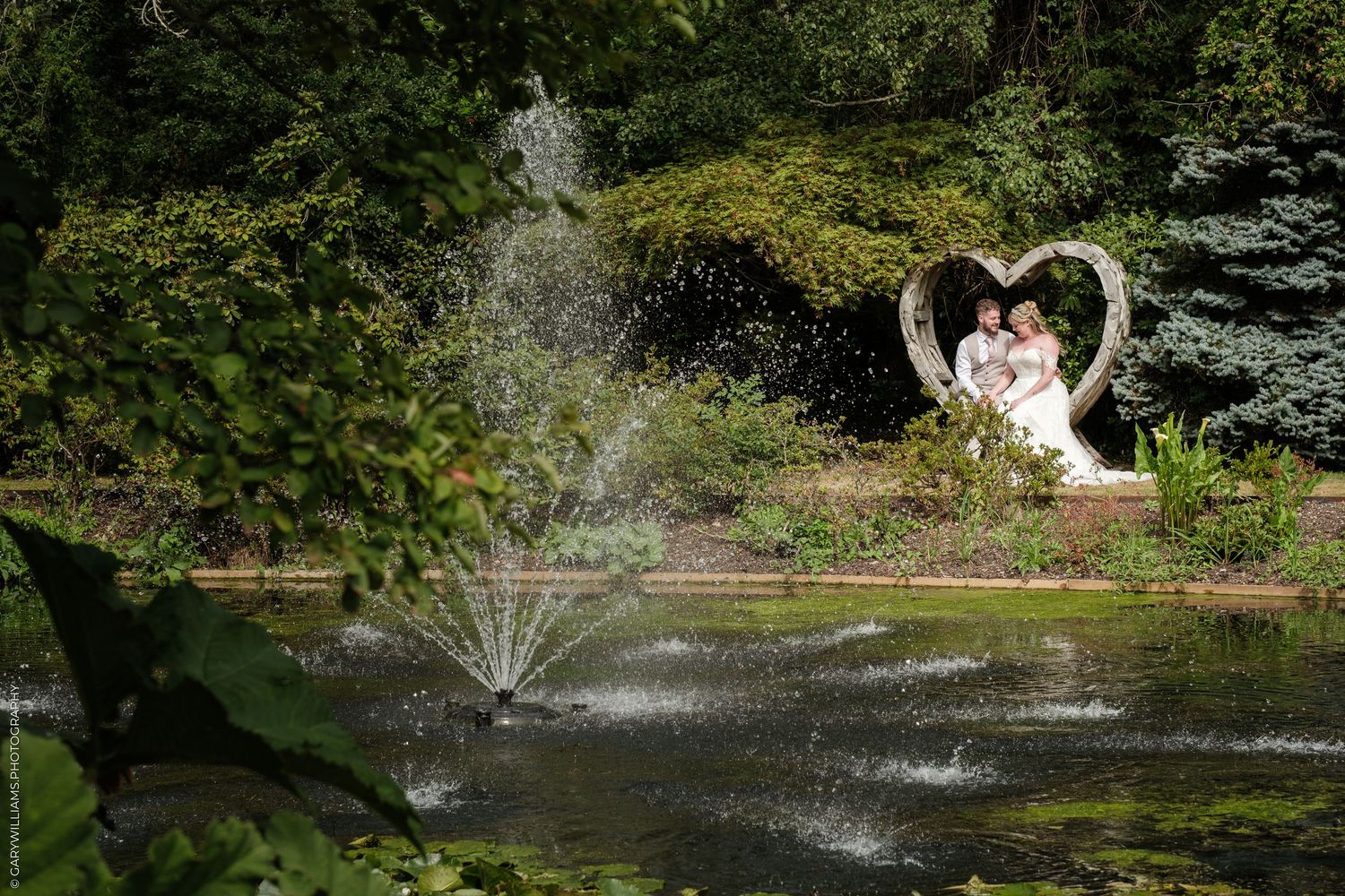 Bride and groom sitting in heart sculpture by a garden pond with fountain surrounded by lush greenery.