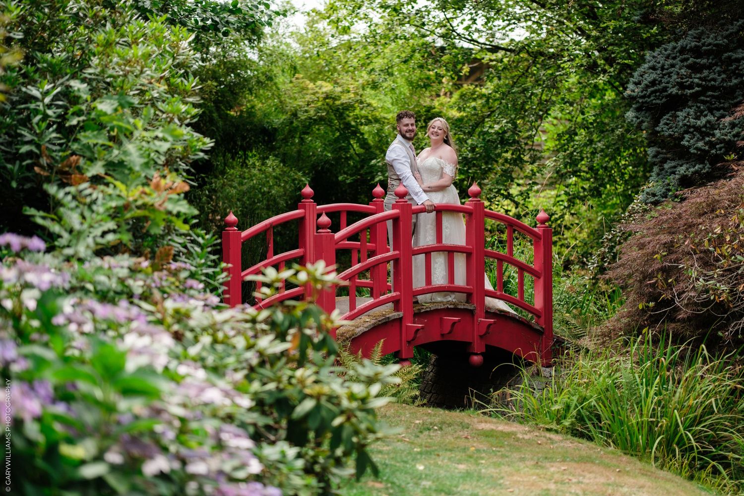 A couple in wedding attire poses on a red Japanese bridge surrounded by lush greenery in a romantic garden setting.