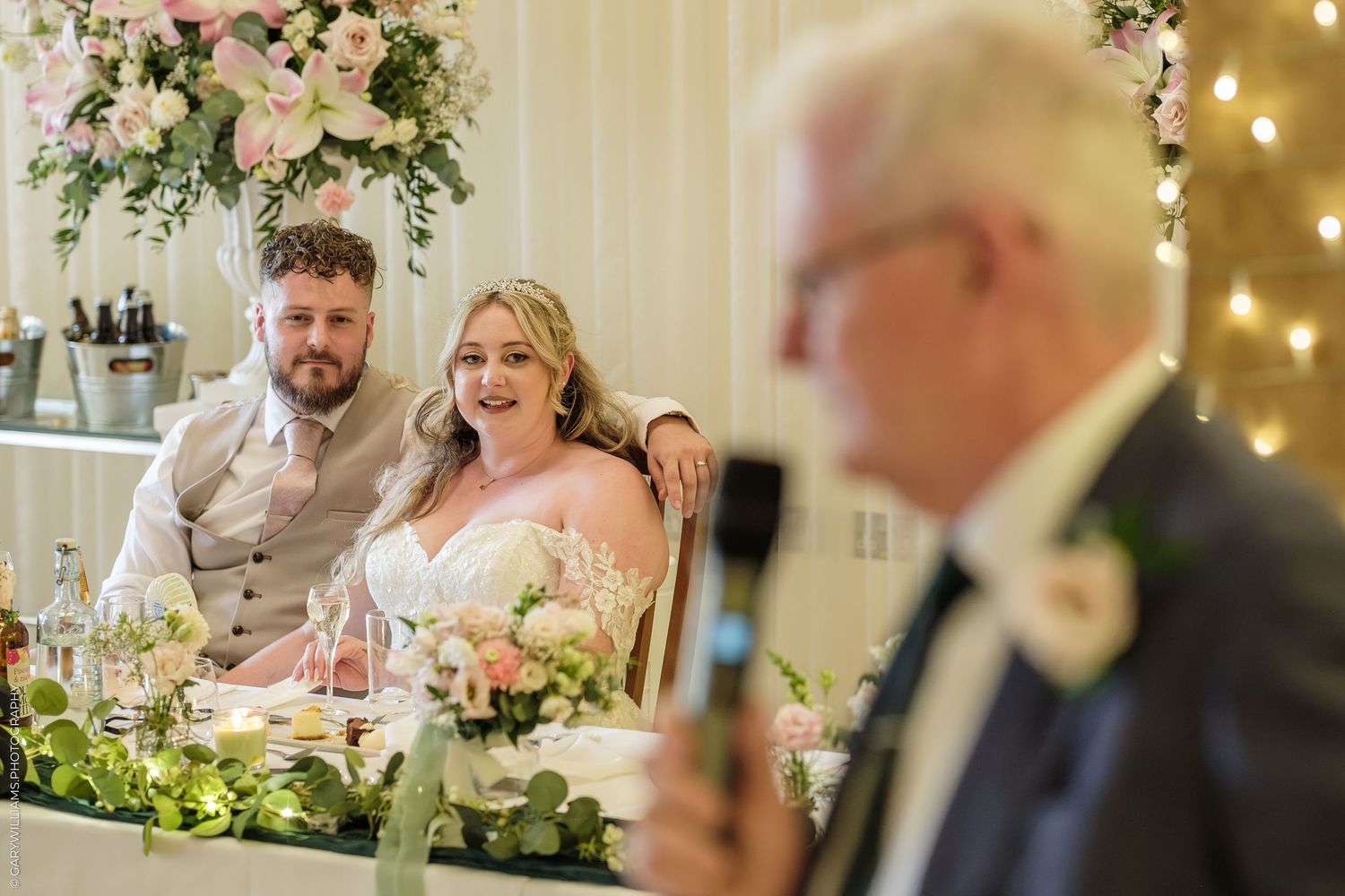 Bride and groom smiling at head table during wedding reception speech, with floral arch backdrop and fairy lights.
