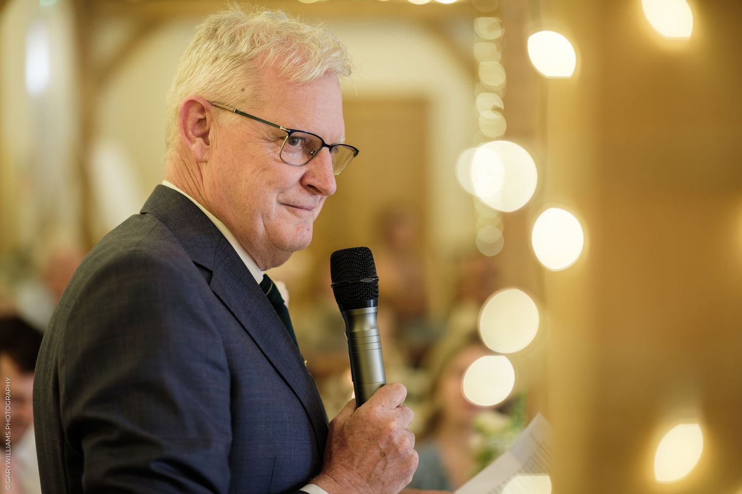 Elderly man in blue suit speaking into microphone at formal event with bokeh lights in background.