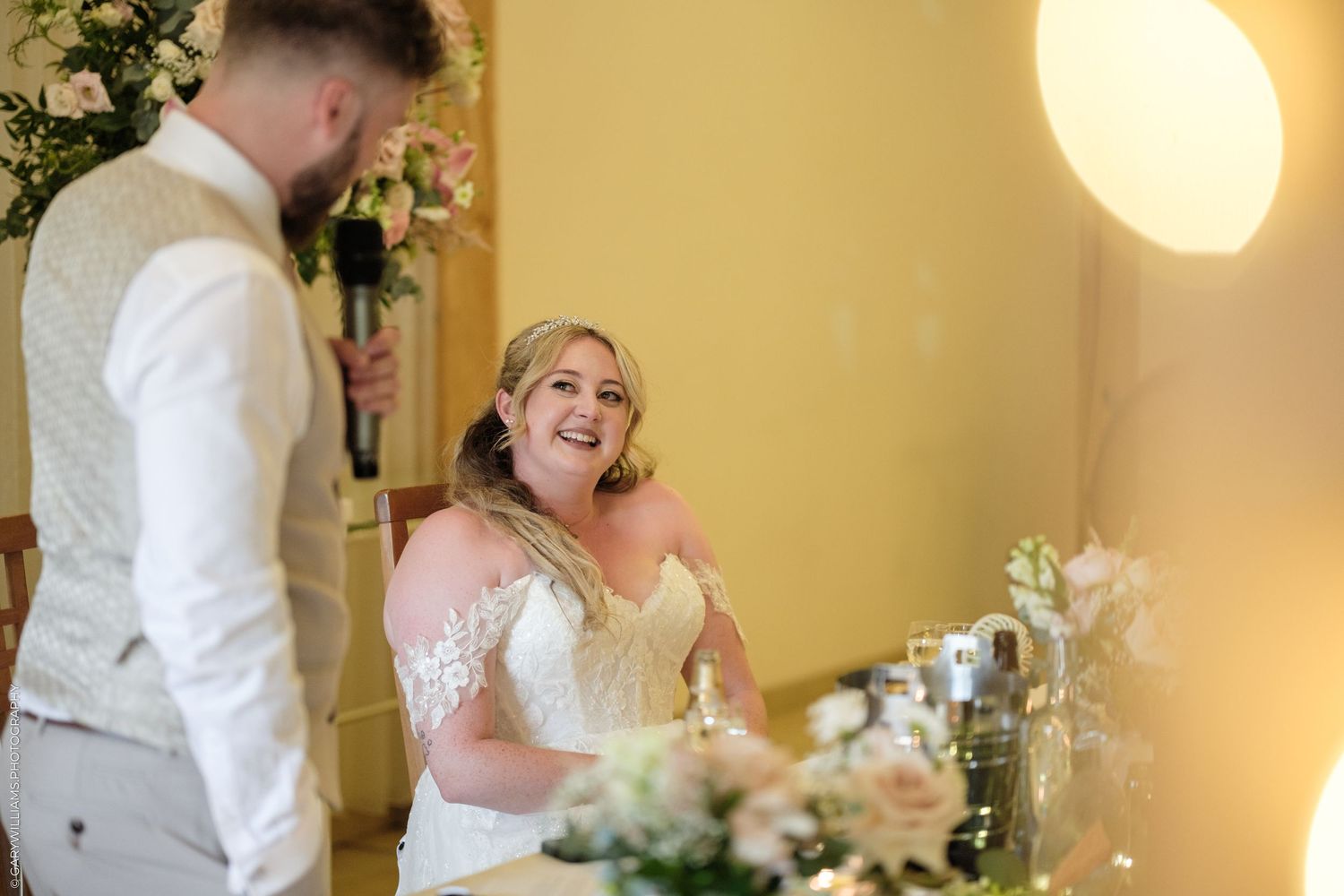 Bride in lace wedding dress laughing joyfully at groom during wedding reception at elegant venue.
