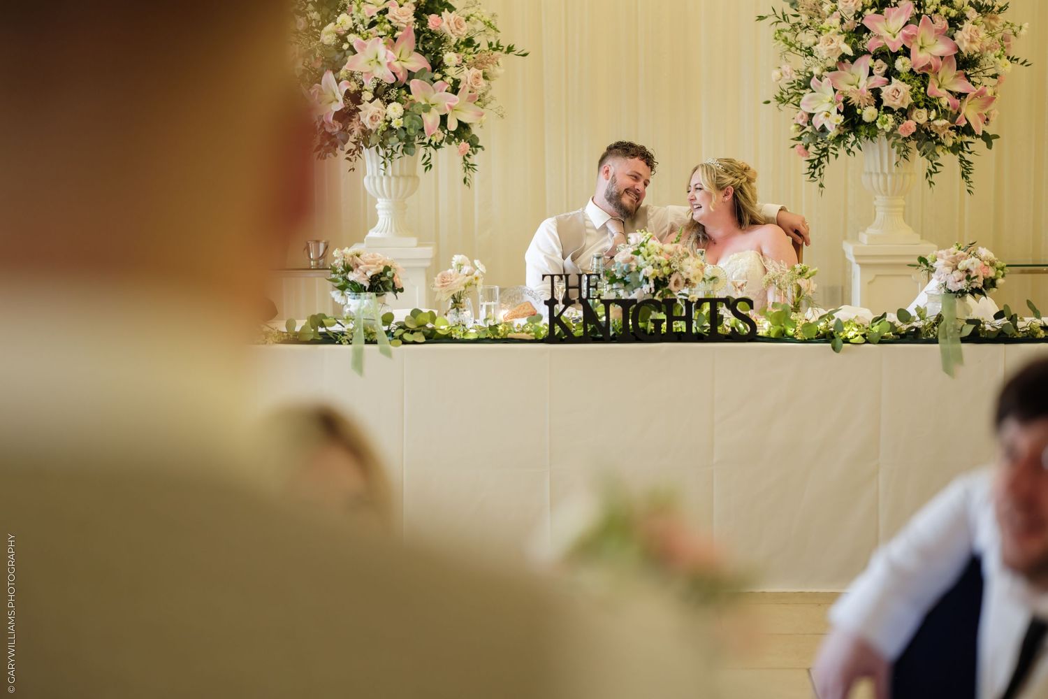 Bride and groom laughing together at wedding reception sweetheart table adorned with pink floral arrangements.