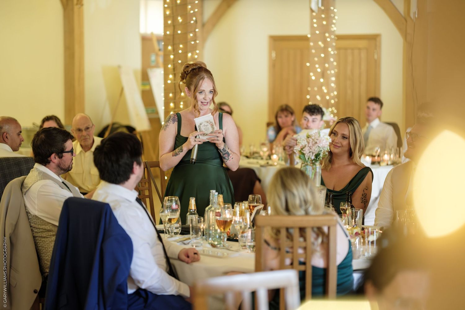 A bridesmaid in a green dress gives a heartfelt speech at an elegant wedding reception with fairy lights.