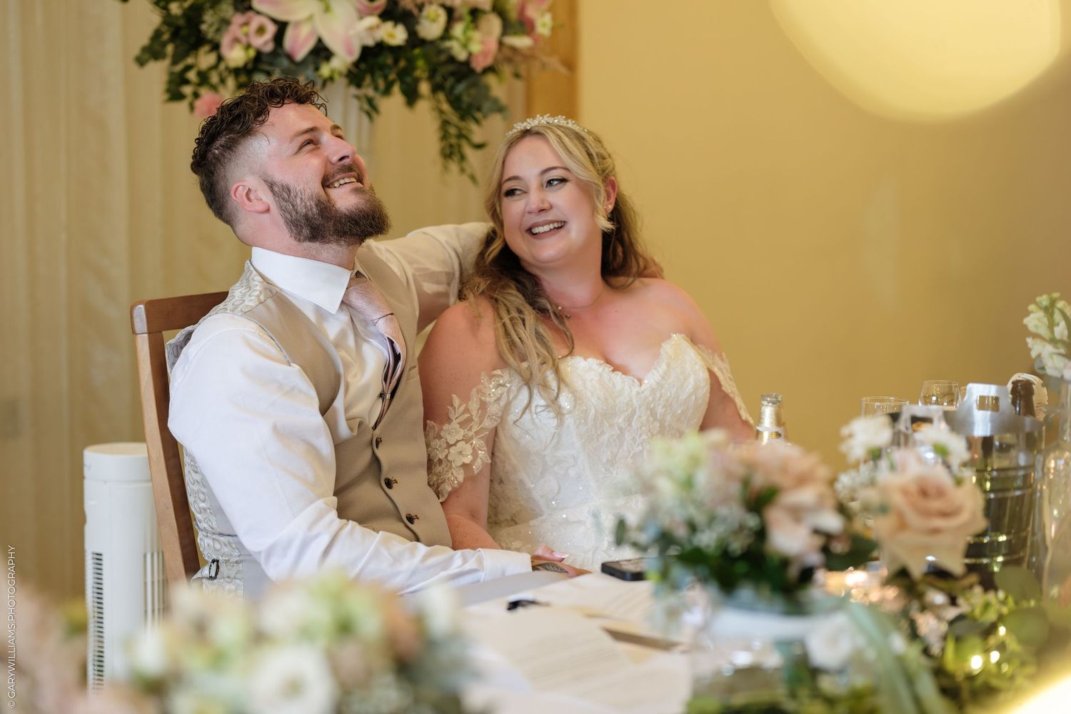 Happy bride and groom laughing at wedding reception table, surrounded by elegant floral arrangements.