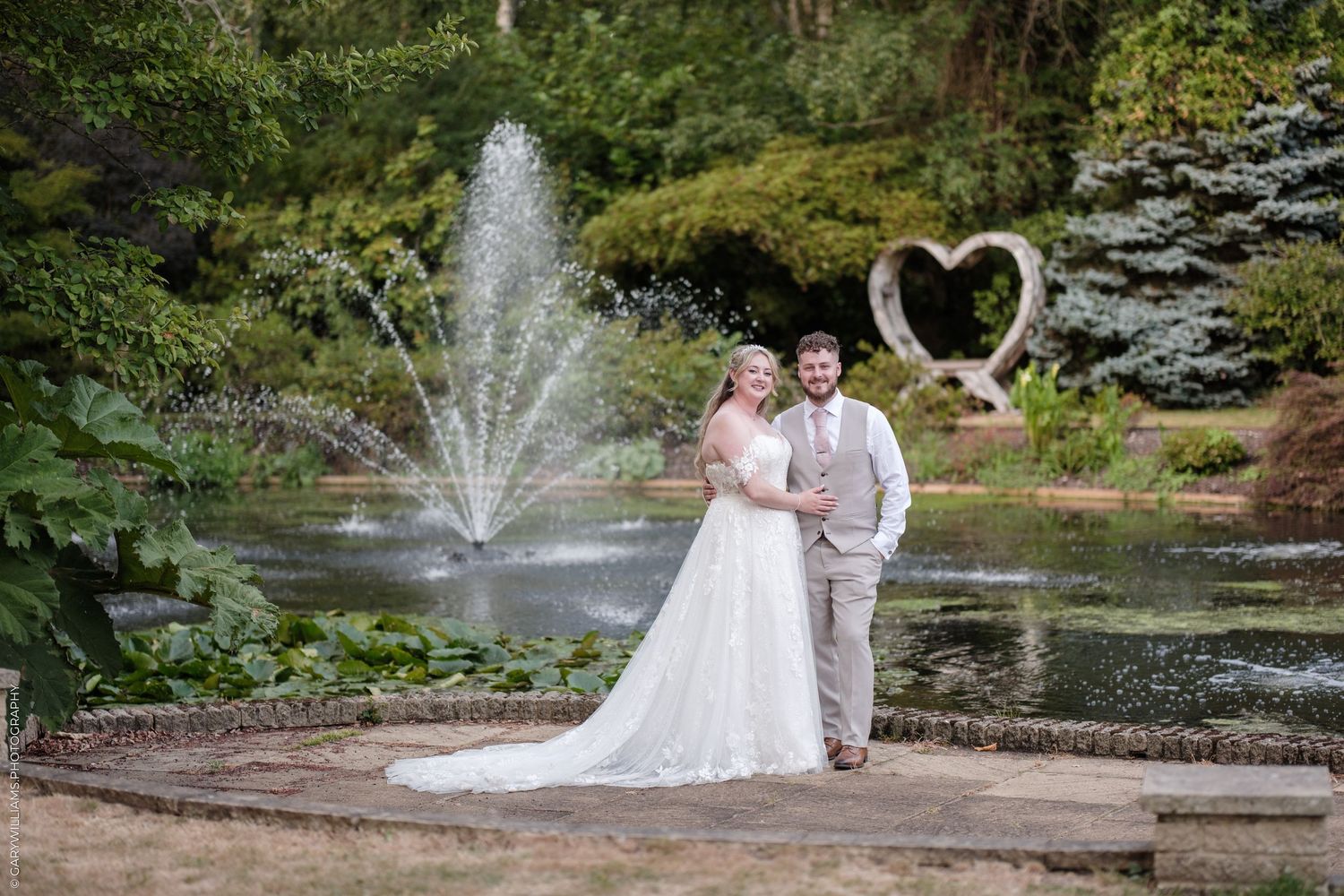Bride and groom pose by a tranquil pond with a fountain and heart sculpture in a lush garden wedding venue.