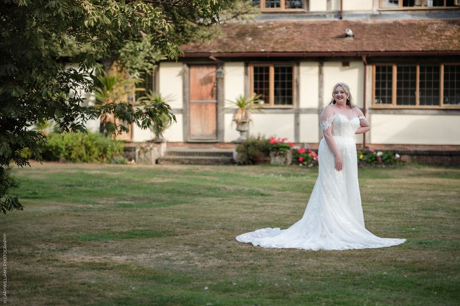 Bride in white lace wedding gown with train standing on green lawn before a rustic manor house.