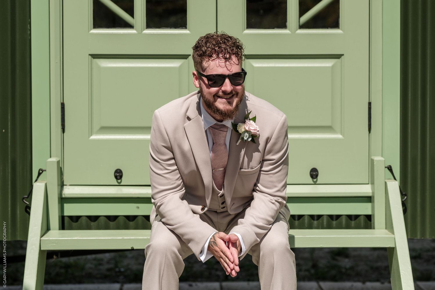 Smiling groom in tan suit with boutonniere sits on bench before green door on sunny wedding day.