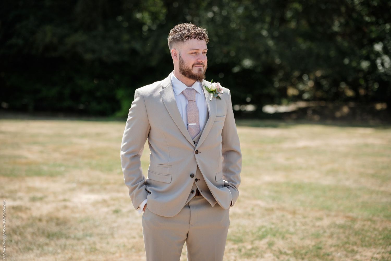 A groom in a tan suit with a floral boutonniere stands outdoors on a sunny day at a wedding venue.