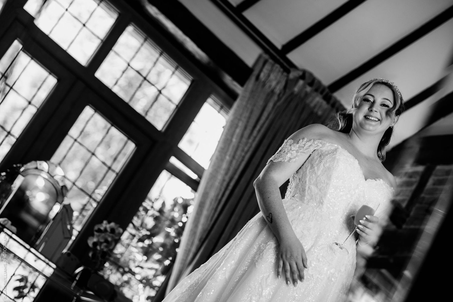 Black and white photo of a smiling bride in a lace wedding dress standing by large windows on her wedding day.