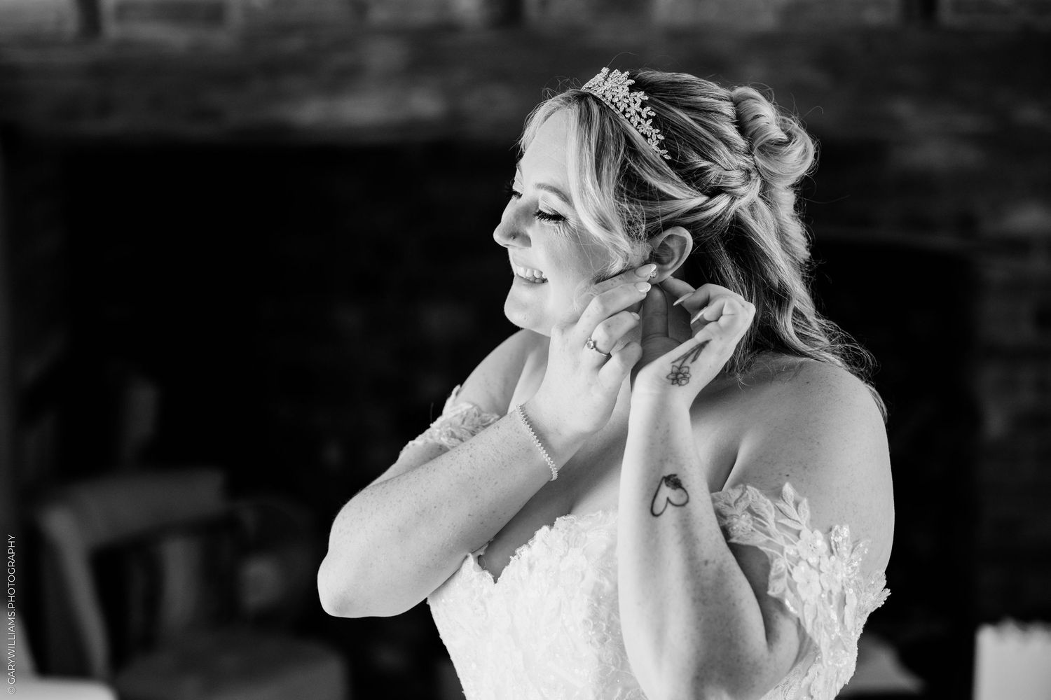Bride in lace gown adjusting earring in black and white wedding photo with tiara and floral tattoo.