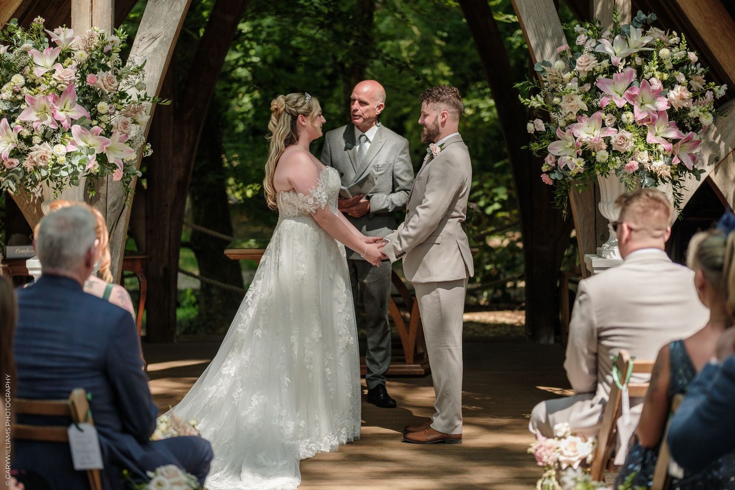 Bride and groom exchange vows at an outdoor woodland wedding ceremony adorned with pink and white floral arrangements.