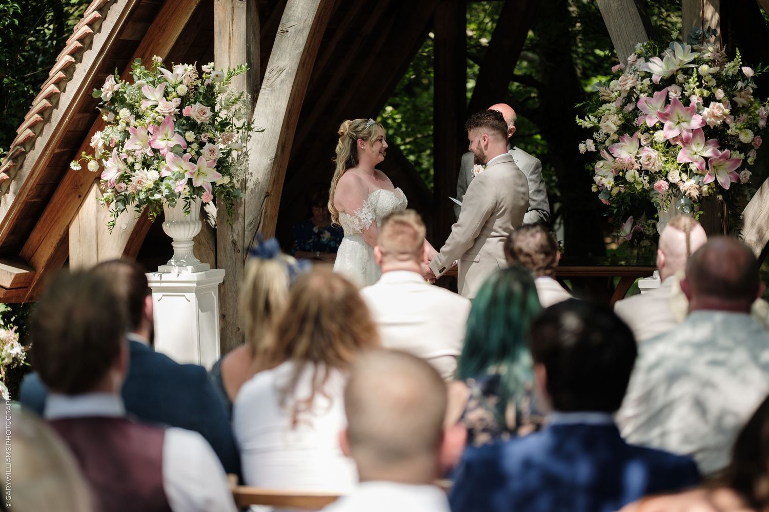 Bride and groom exchange vows at an outdoor wedding ceremony surrounded by lush floral arrangements and seated guests.
