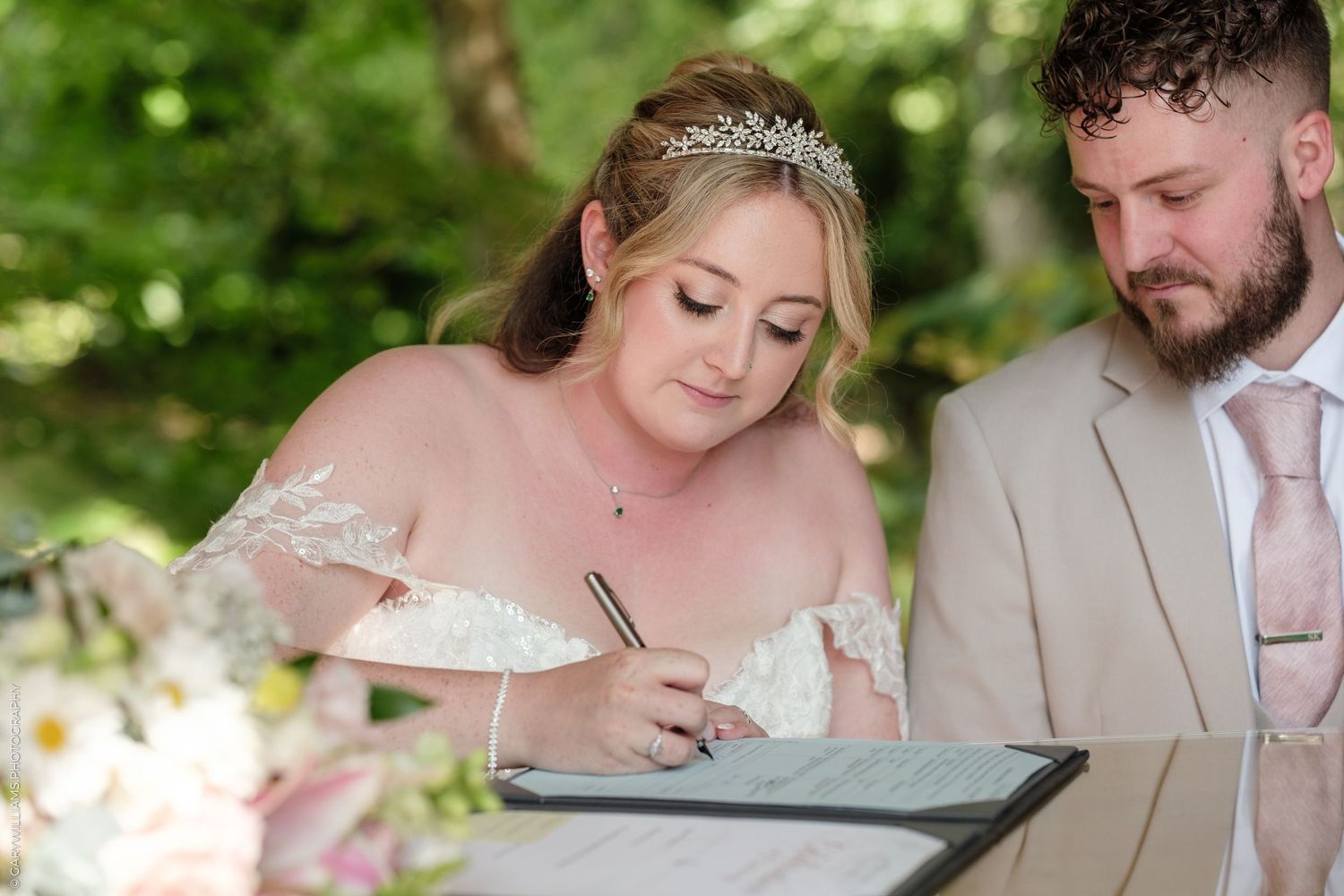 Bride in lace gown and tiara signing wedding register alongside bearded groom in tan suit outdoors.