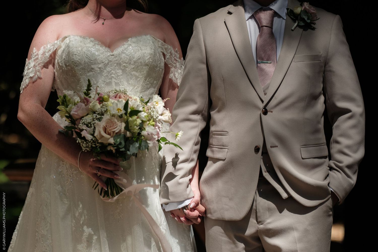 Bride in lace off-shoulder gown holding floral bouquet stands beside groom in beige suit, holding hands at wedding ceremony.