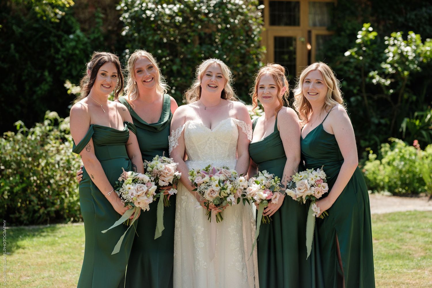 Bride in white gown with four bridesmaids in emerald green dresses holding bouquets outdoors.
