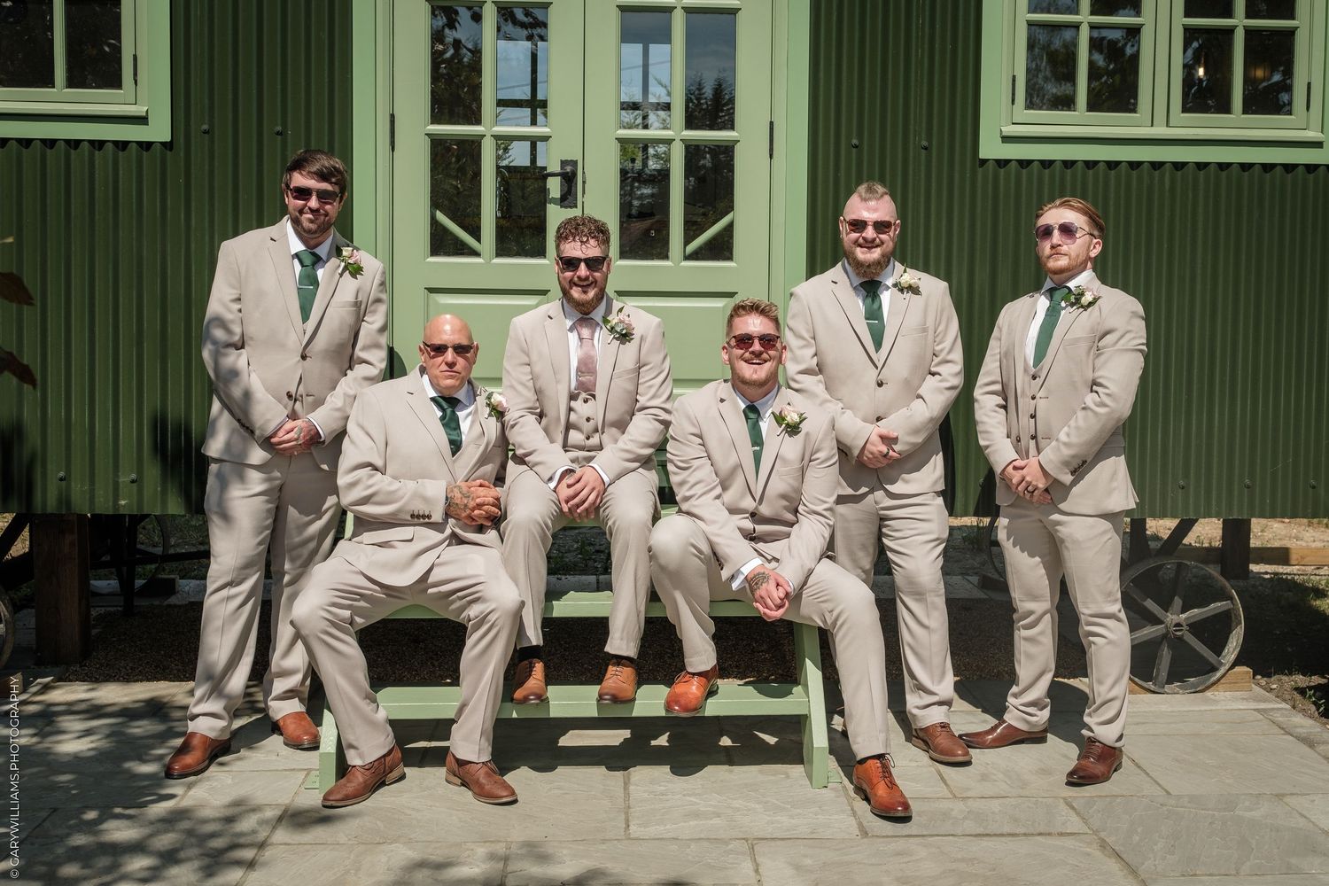 Six groomsmen in matching tan suits with green ties pose outdoors in front of a green building on a sunny wedding day.