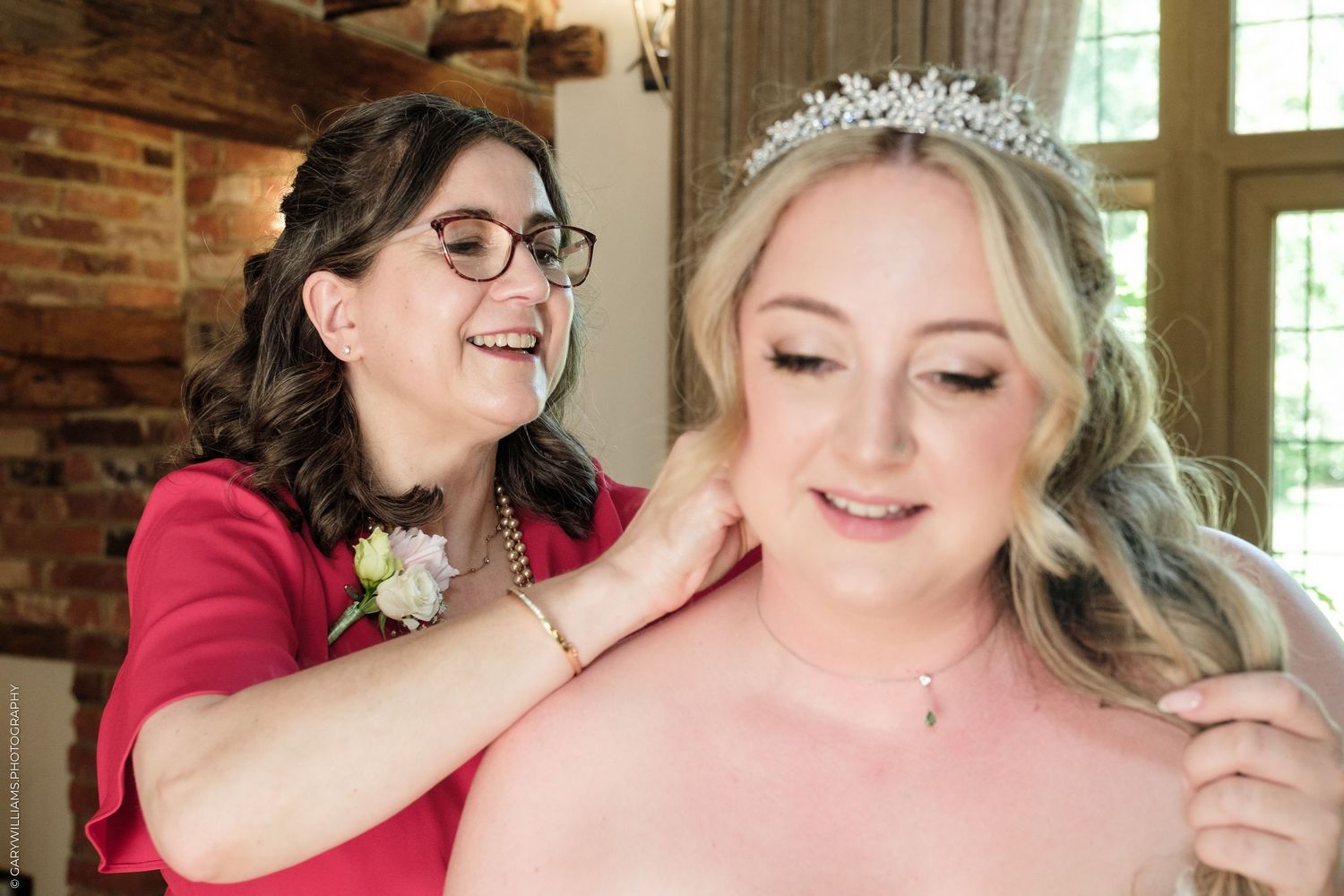 Mother helping bride with necklace, bride wearing tiara, both smiling, wedding preparation in rustic venue.