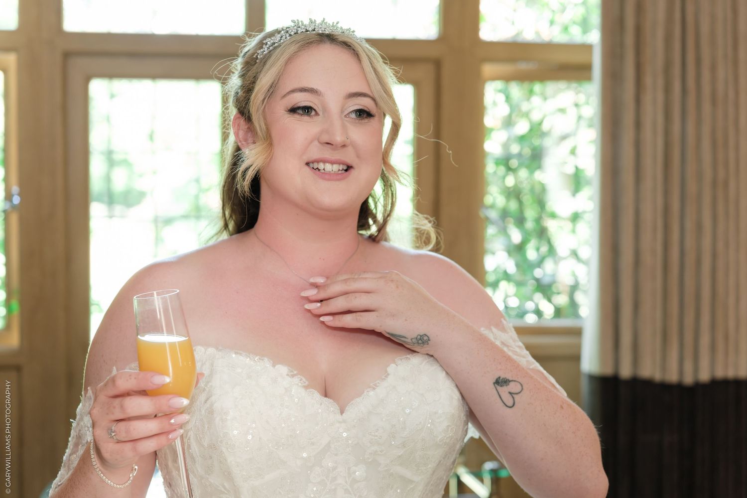 Smiling bride in white lace gown holding champagne glass, wearing tiara and showing heart tattoo on arm.
