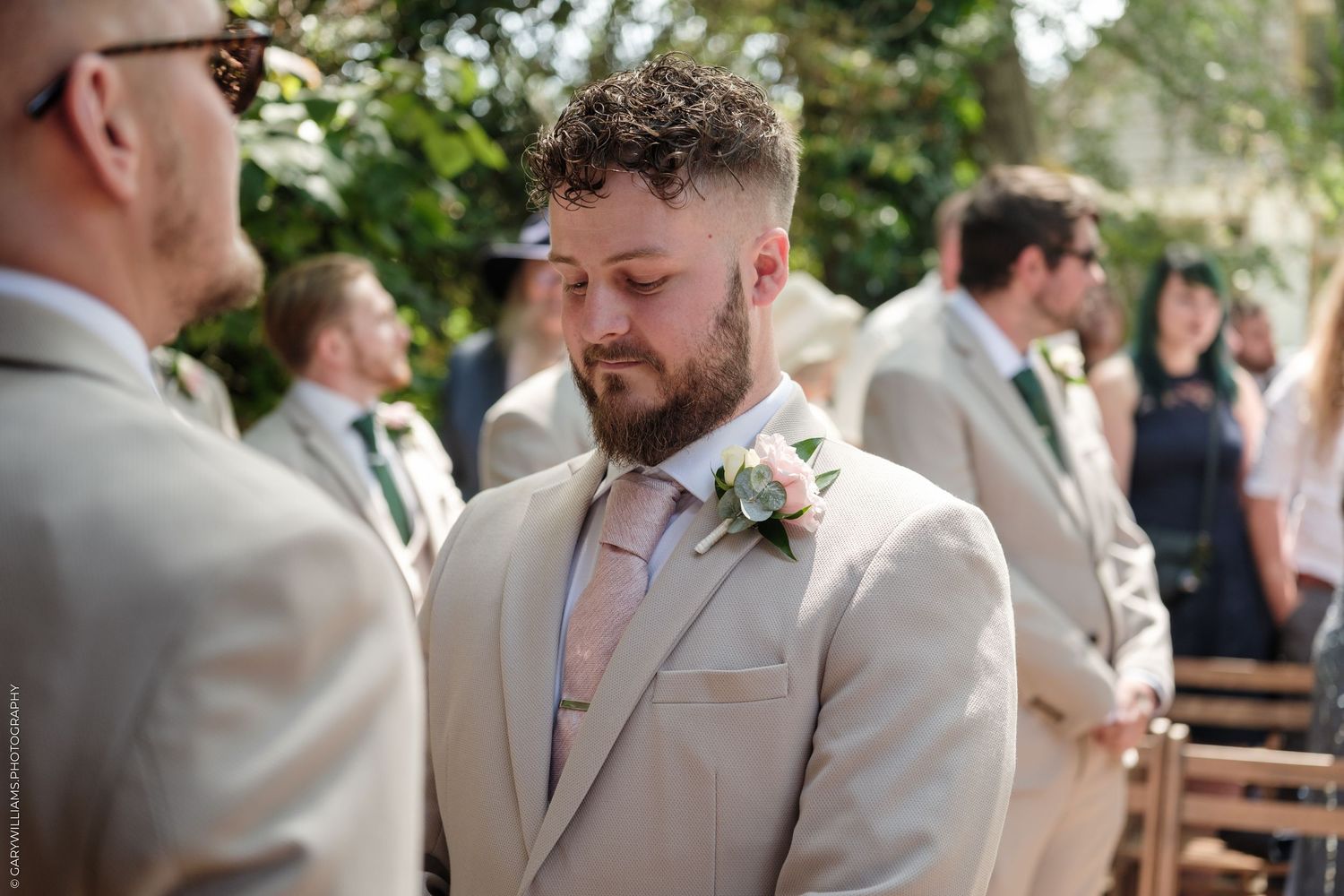 Groom in beige suit with pink boutonniere stands at outdoor wedding ceremony, eyes downcast, groomsmen visible behind him.