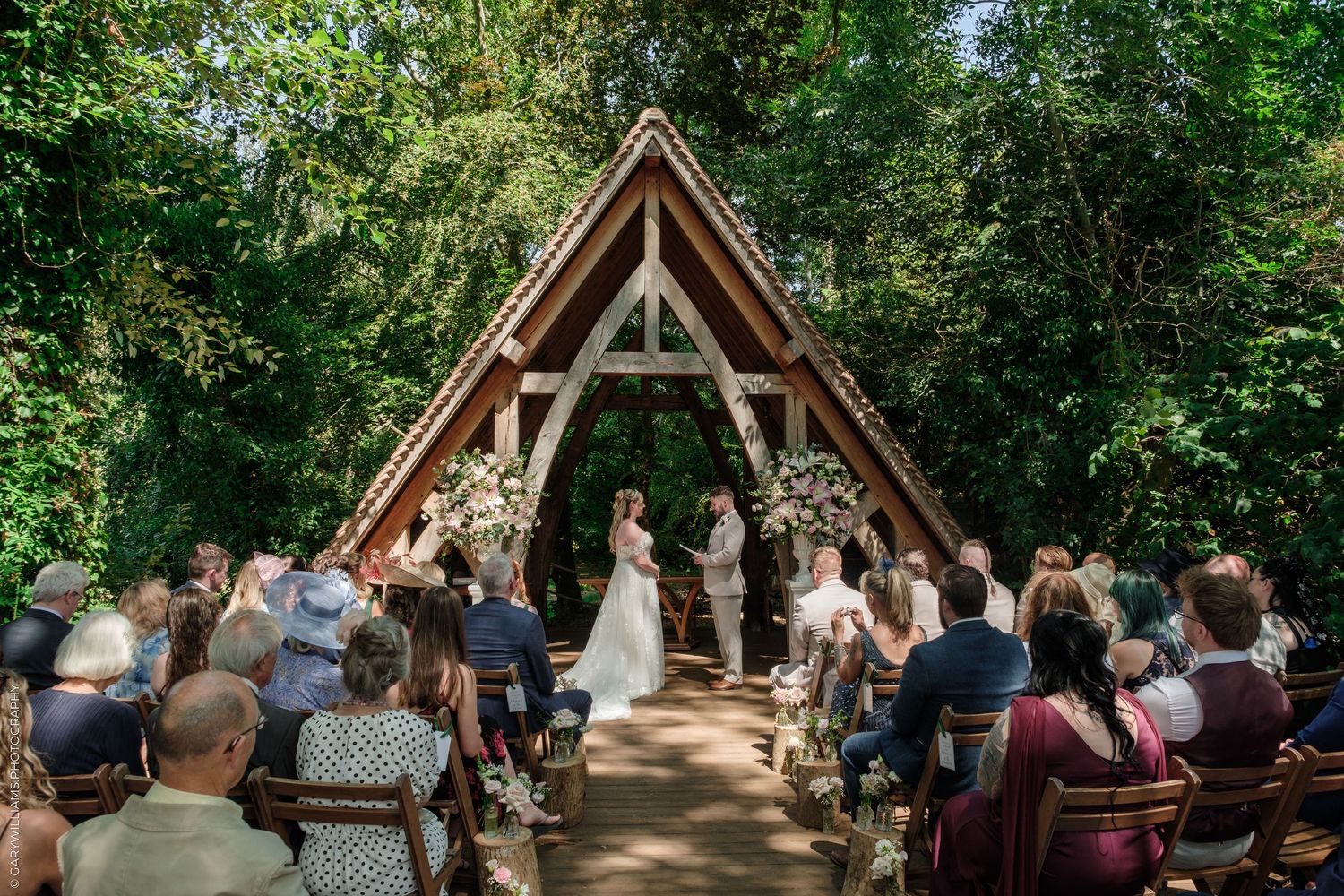 Outdoor wedding ceremony in a lush forest setting with an A-frame wooden chapel, guests seated along an aisle.
