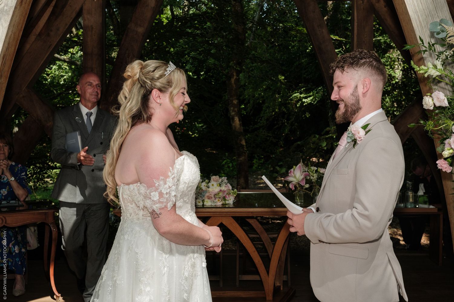 Bride and groom exchanging vows at an outdoor woodland wedding ceremony, officiant visible in background.