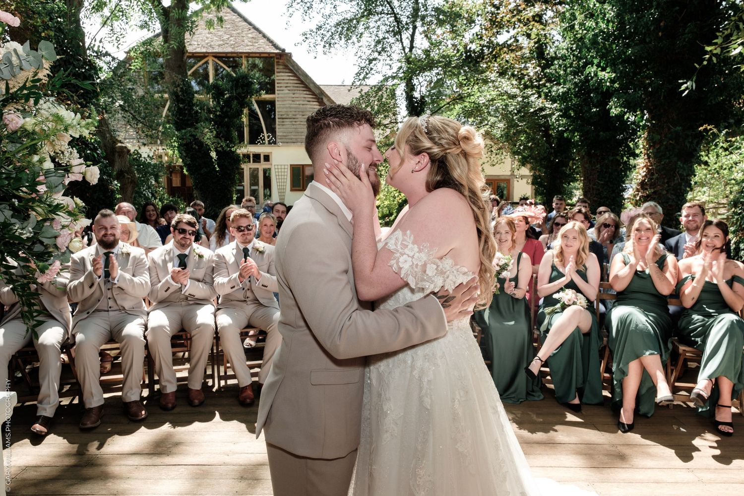 Newlyweds share their first kiss at an outdoor wedding ceremony surrounded by applauding guests.