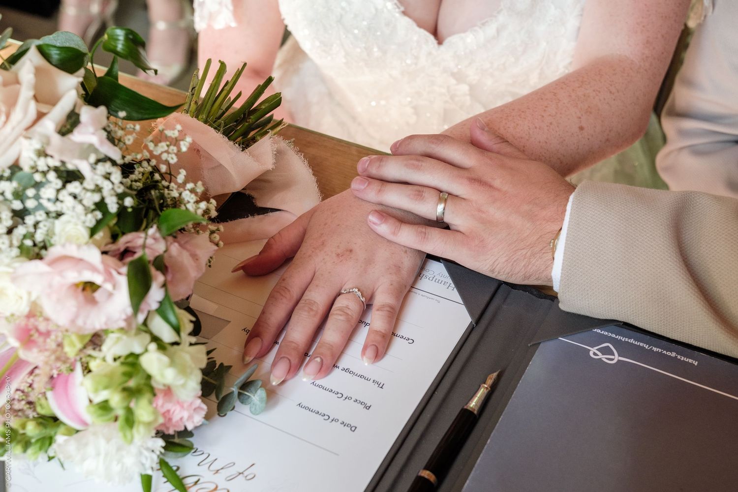 Bride and groom display wedding rings while signing marriage certificate beside pink floral bouquet.