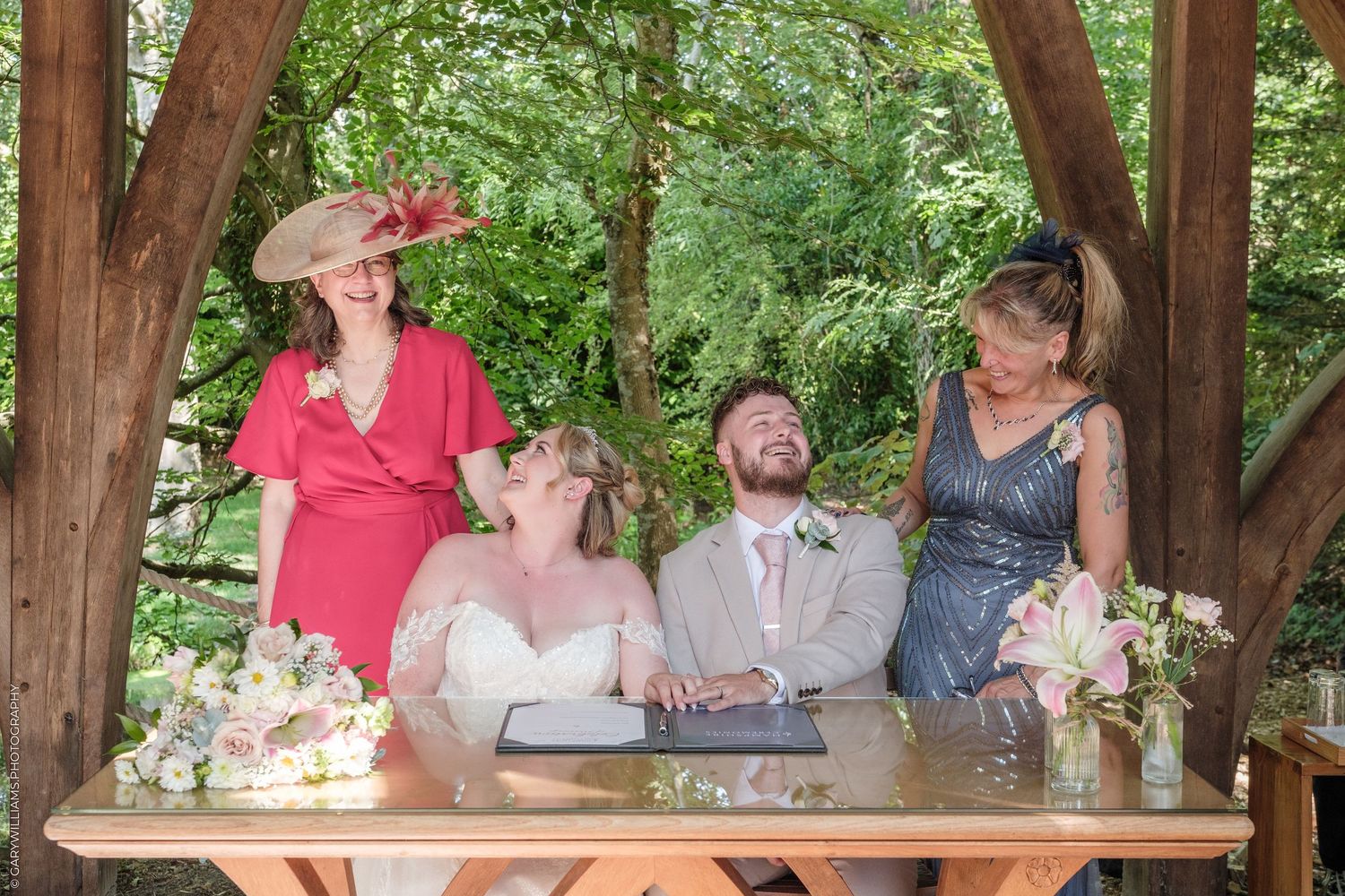 Bride and groom sign wedding documents at outdoor gazebo table, joined by two smiling women in pink and grey dresses.