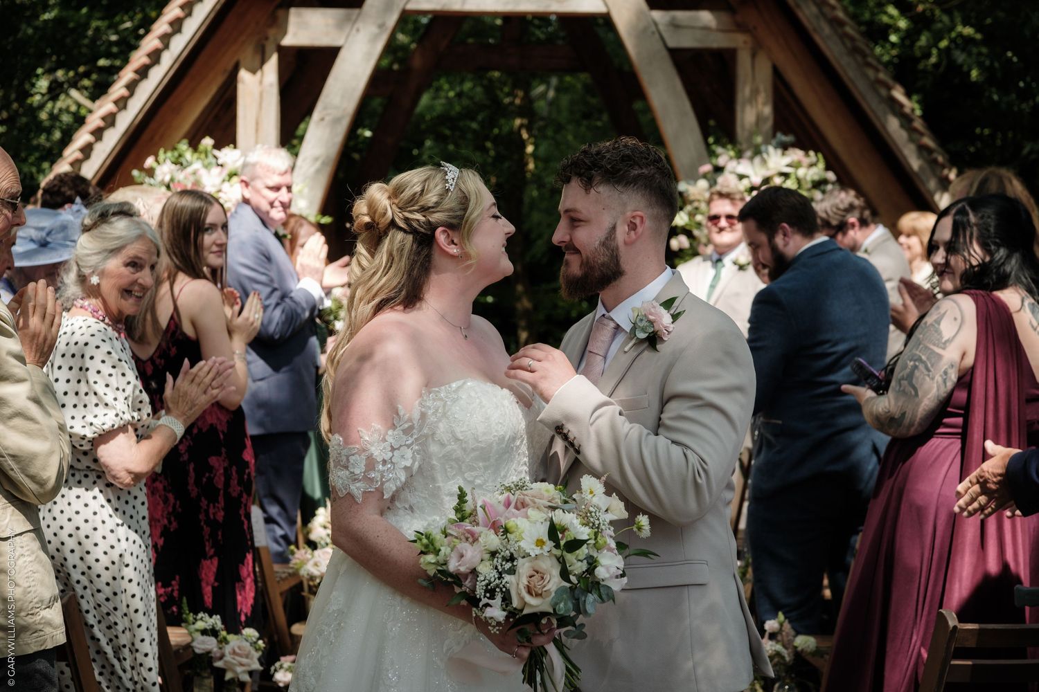 Bride and groom share a joyful moment at outdoor wedding ceremony surrounded by cheering guests and floral arch.