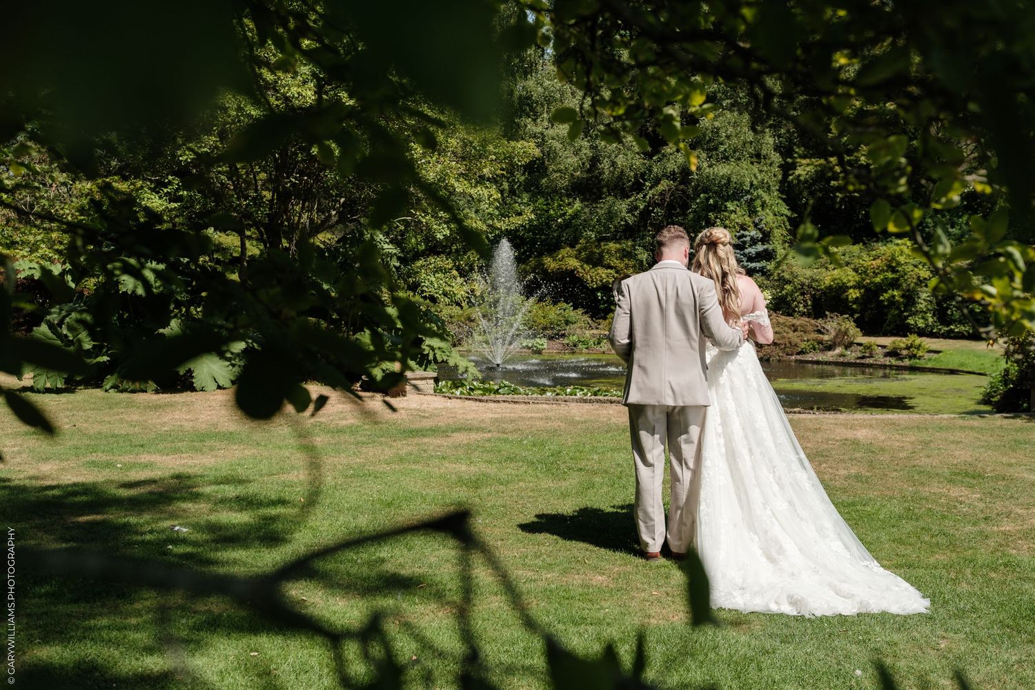Bride and groom walk hand in hand through a lush green garden with a fountain backdrop on their wedding day.