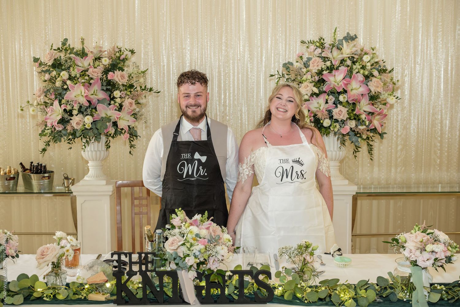 Newlyweds wearing Mr. and Mrs. aprons smile together at their wedding reception table adorned with pink floral arrangements.