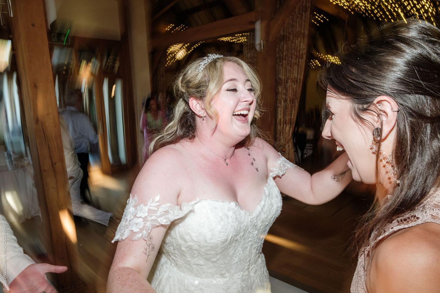 Joyful bride in lace wedding dress laughs with bridesmaid inside rustic barn wedding venue.