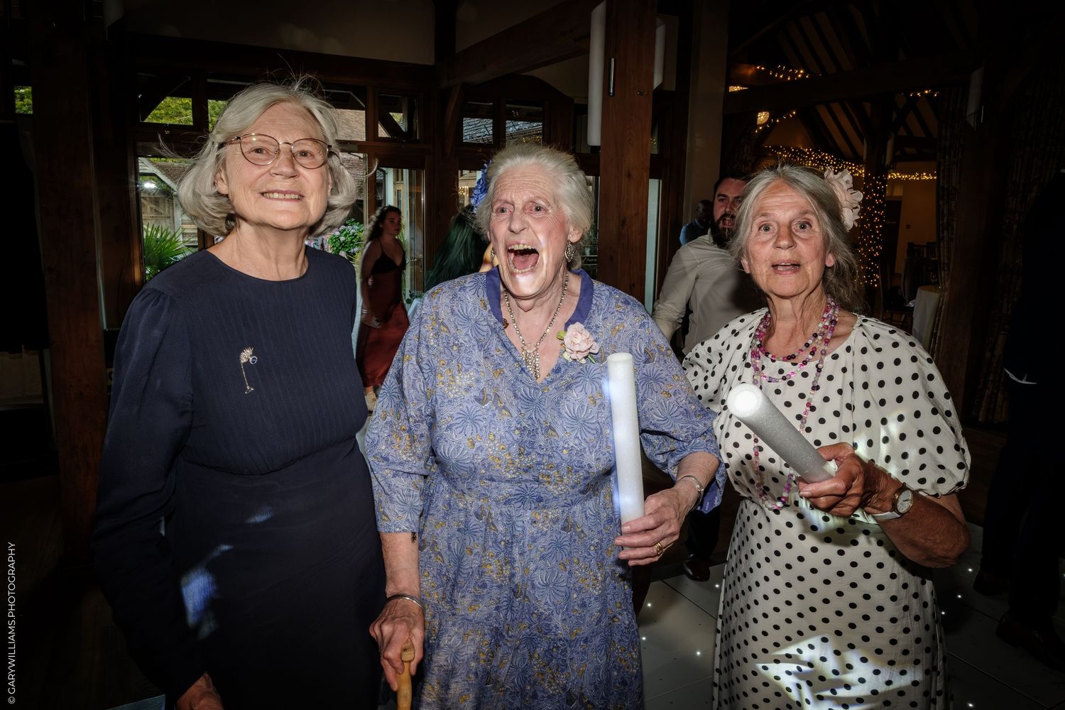 Three elderly women smiling and laughing together at a celebration event indoors.