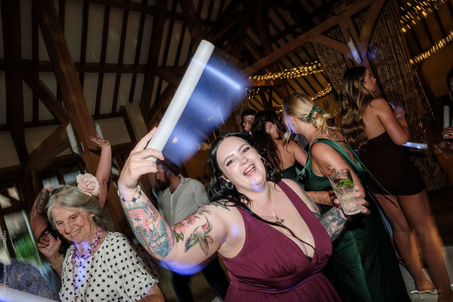 Joyful wedding guest with tattoos dances energetically holding a foam tube at barn reception.