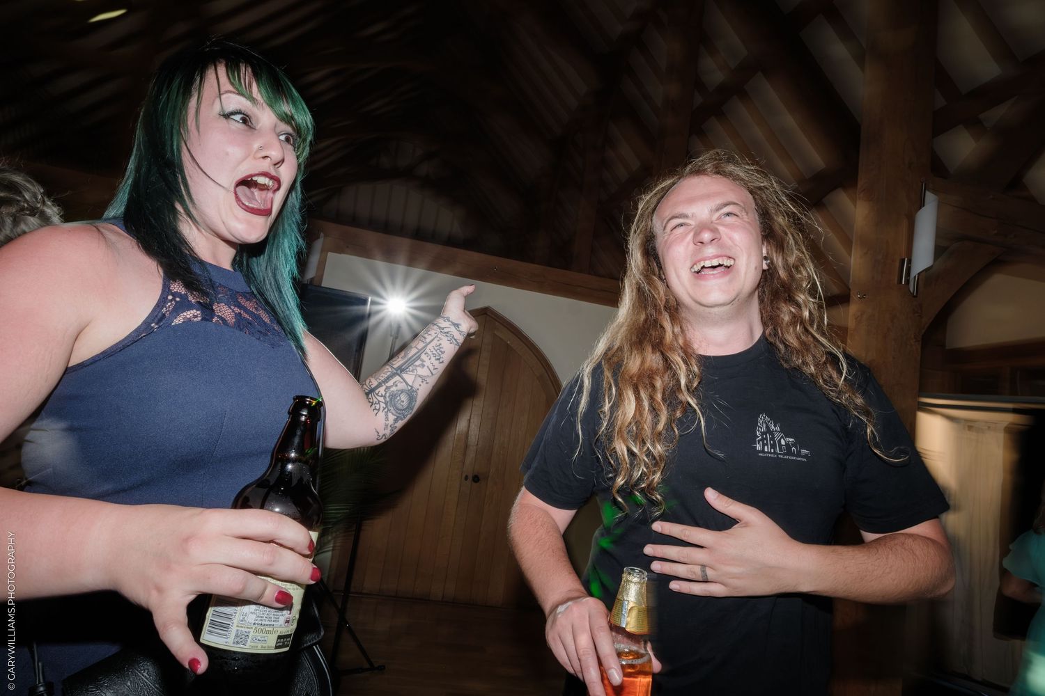 Two women laughing and holding beer bottles at an indoor party with wooden ceiling.