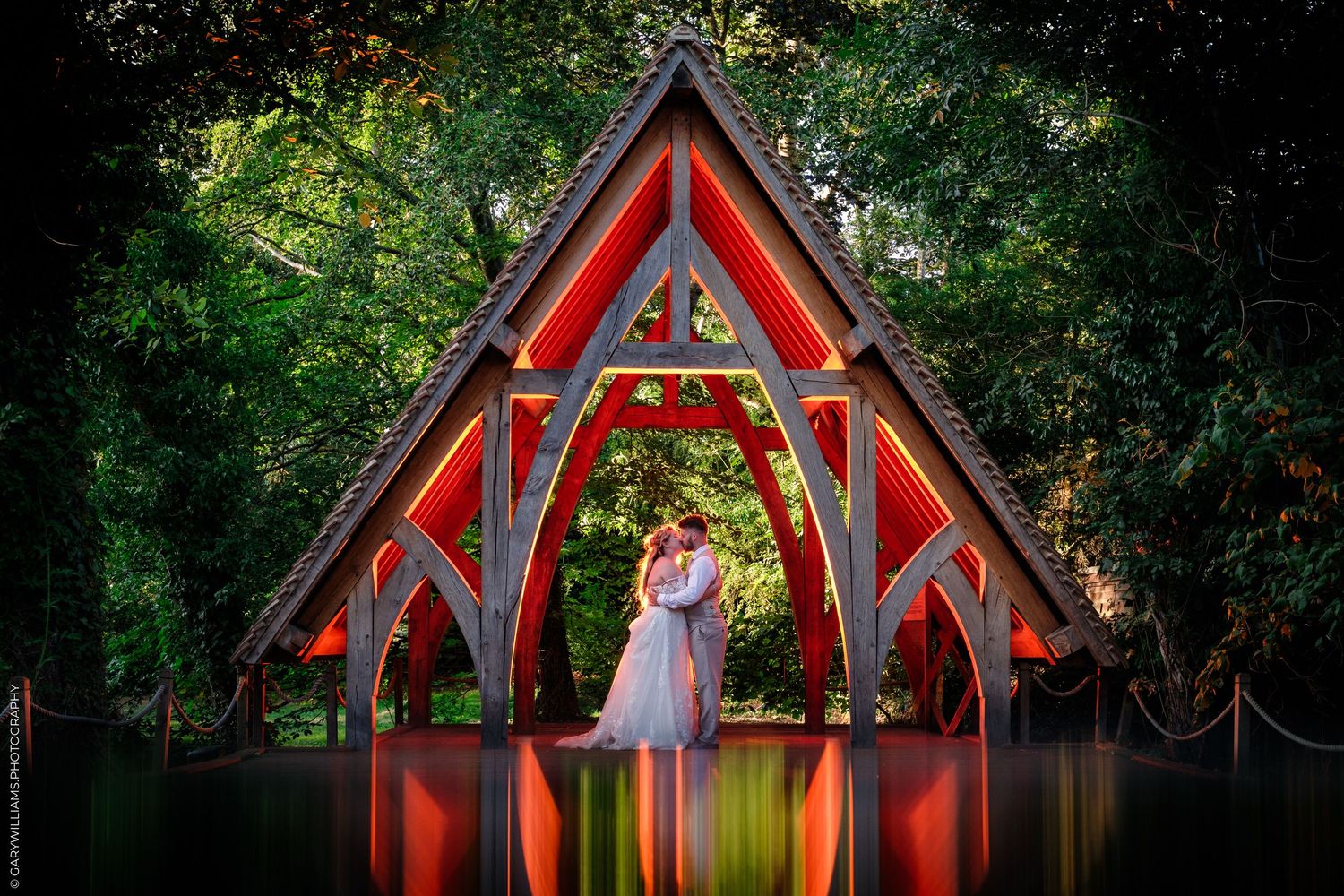 Couple embracing under a red illuminated wooden arch pavilion surrounded by lush green trees at dusk.