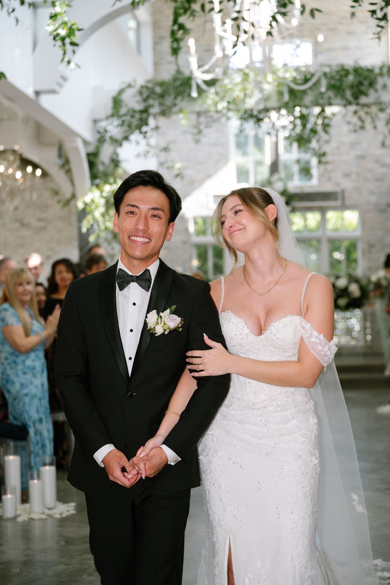 Bride and groom walking down the aisle, smiling joyfully, in an elegant venue with greenery decor.