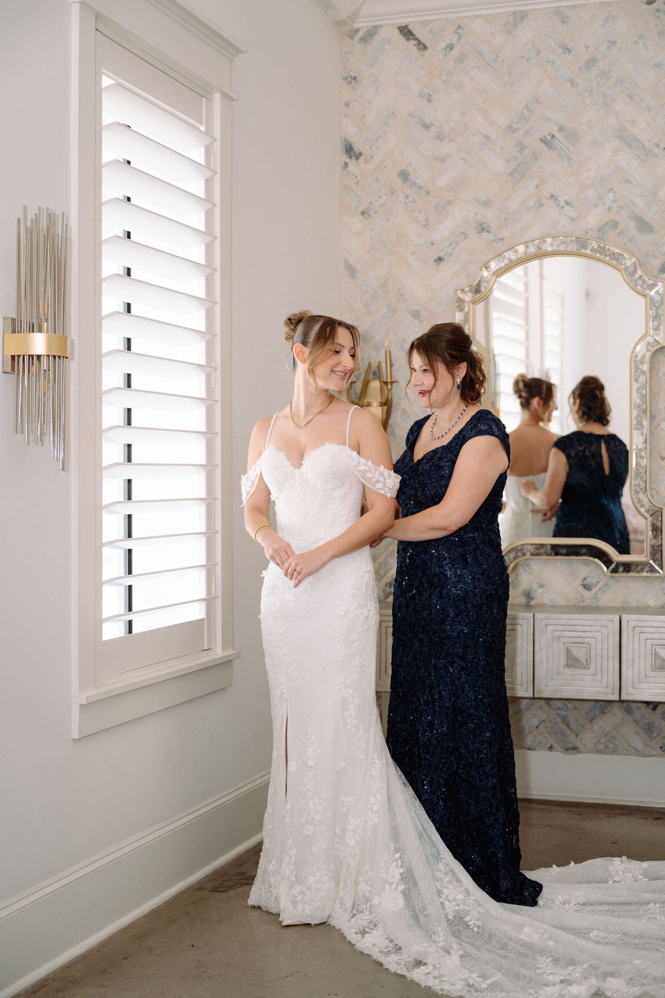 Bride in white lace gown with off-shoulder sleeves stands with woman in navy dress in elegant bridal suite.