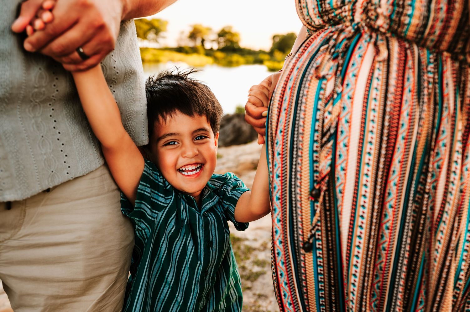 A laughing young boy holds the hands of two adults outdoors near a rocky riverbank at golden hour.