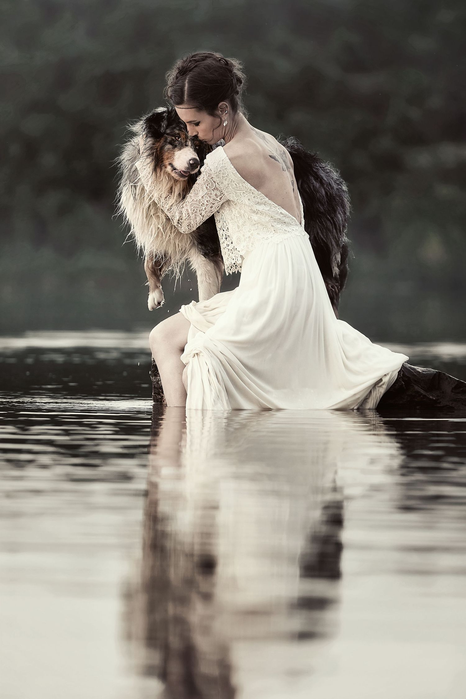 Woman in white lace dress sitting in water, tenderly kissing her Australian Shepherd dog, with calm lake reflections.