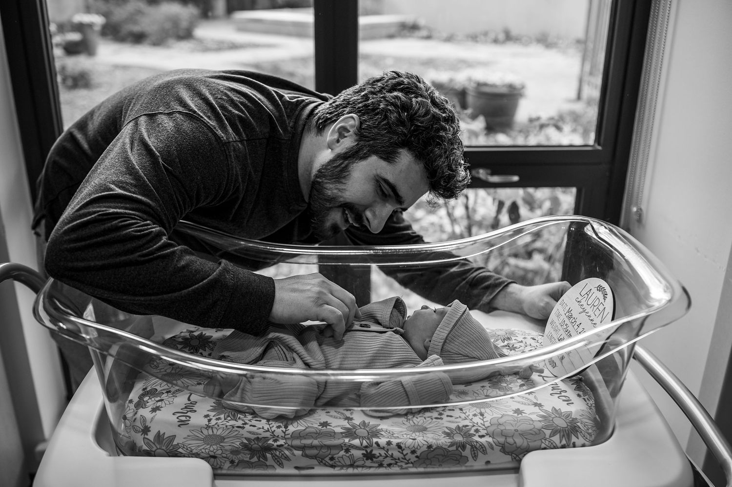 Father tenderly leaning over newborn baby in hospital bassinet in black and white photo.