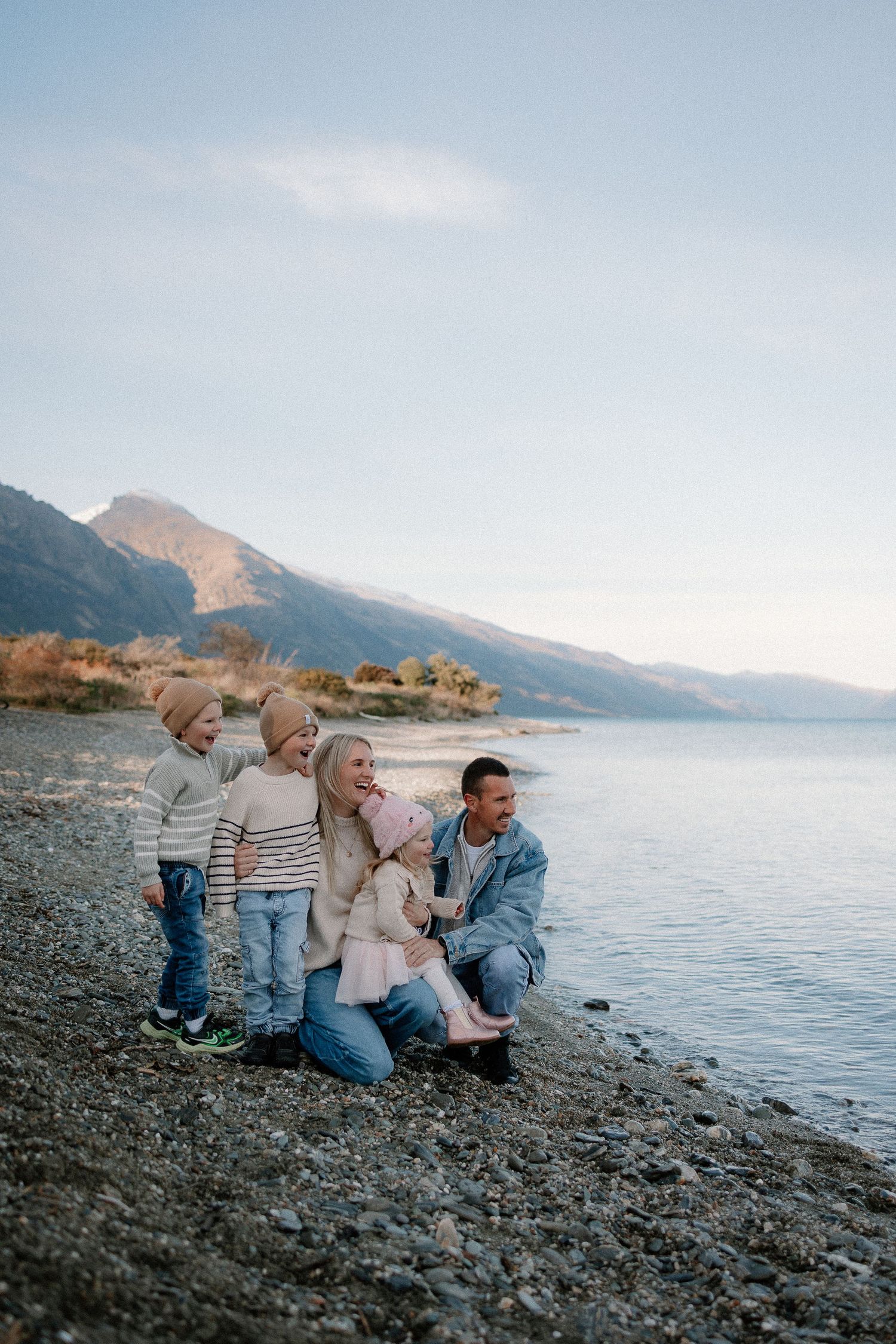 Family of four posing on rocky lakeshore with mountain backdrop on a calm, clear day.