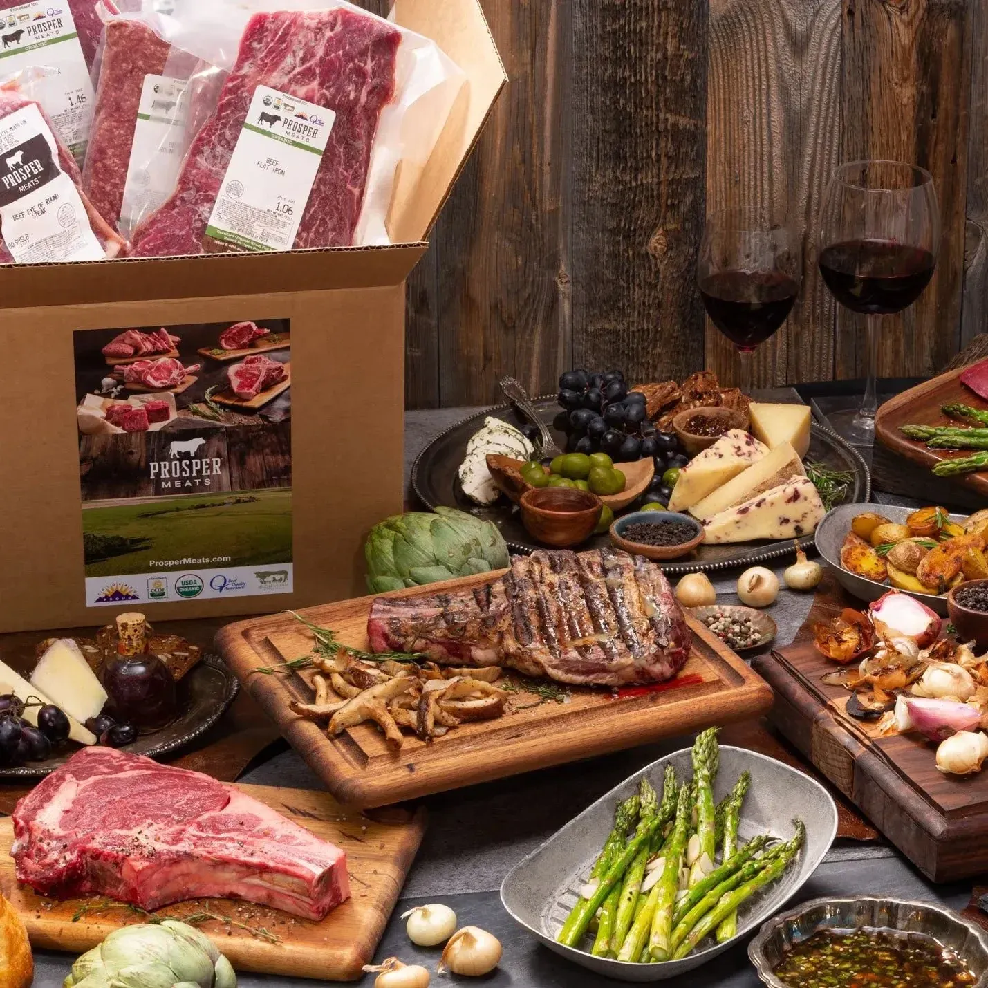 Pasture Prime meat delivery box surrounded by steaks, cheese, grapes, asparagus, and red wine.