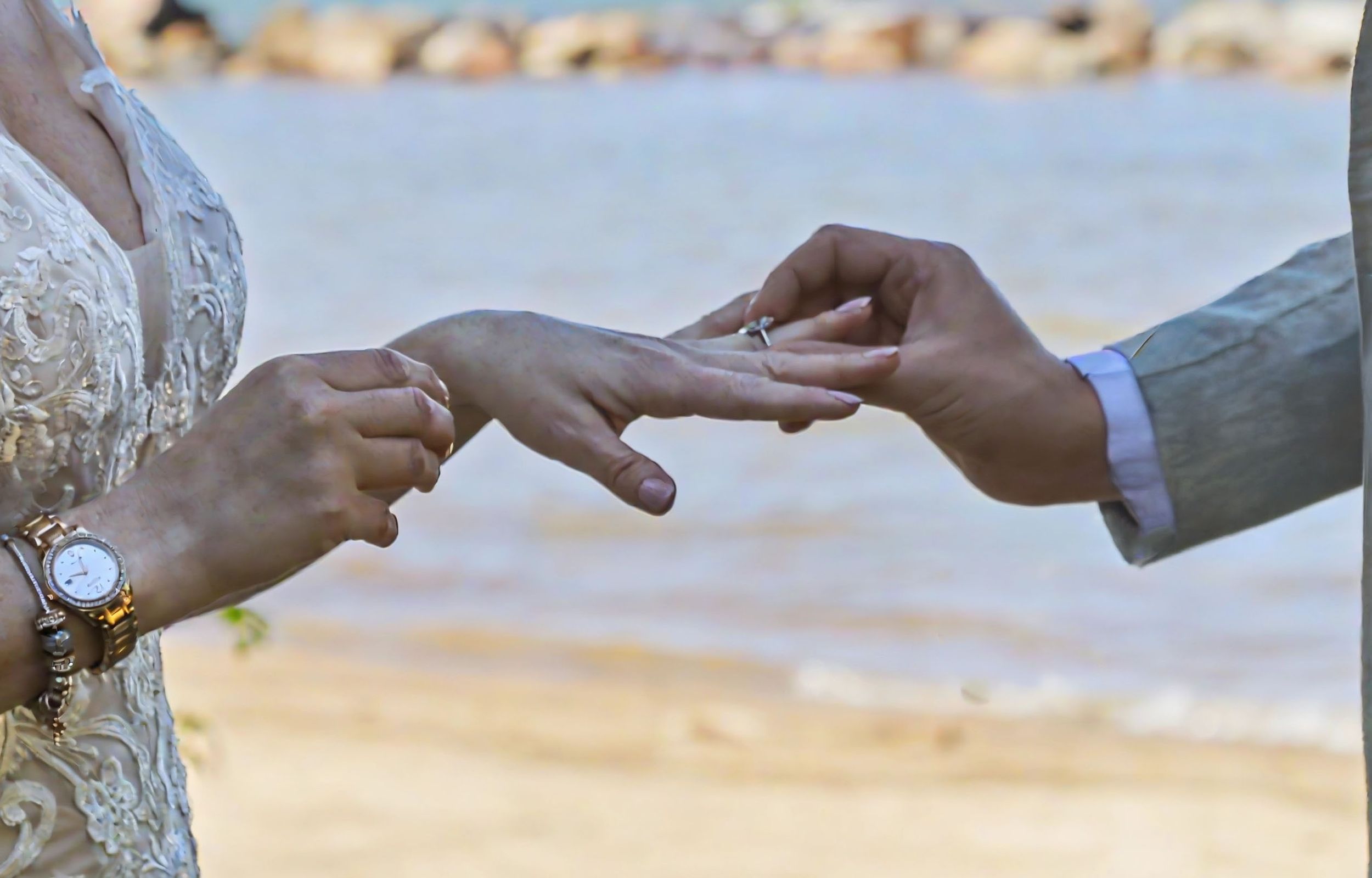 Close-up of a groom placing a wedding ring on the bride's finger during an outdoor beach ceremony.