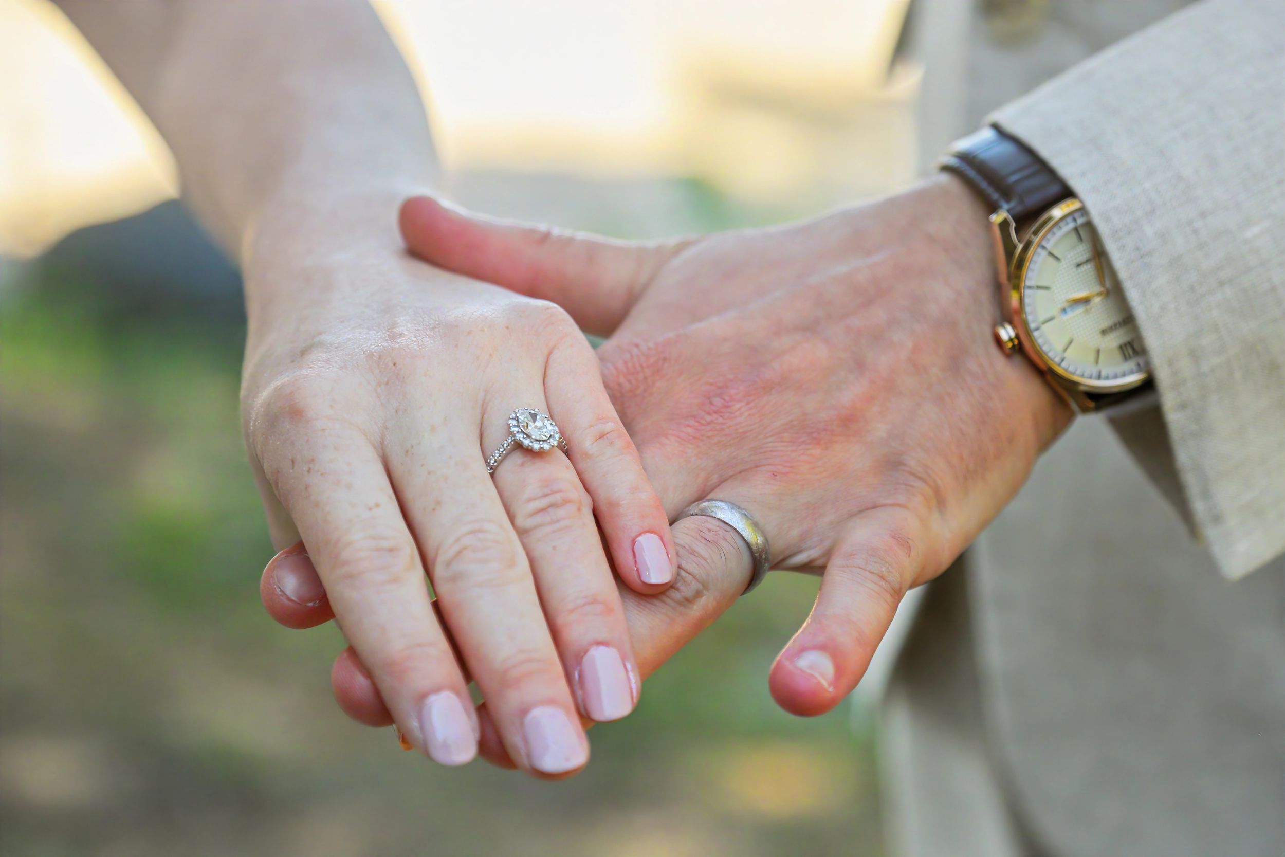 Close-up of a couple holding hands, woman wearing a diamond engagement ring, man wearing a gold watch and wedding band.
